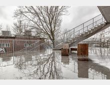 Nasser, spiegelnder Betonboden auf einer offenen Terrasse mit moderner Metalltreppe, Holzbänken und einem Geländer. Im Hintergrund ein Backsteingebäude und kahle Bäume bei bewölktem Himmel.