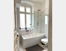 Helles, weiß gefliestes Badezimmer mit Badewanne und integrierter Duschwand aus Glas sowie Regendusche. Neben einem großen, hohen Fenster mit Rundbogen befinden sich eine Fensterbank mit Pflegeprodukten und rechts ein wandhängendes WC.