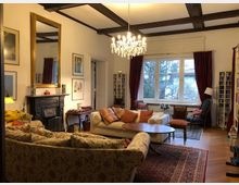 Wohnzimmer mit gemütlicher Einrichtung, bestehend aus gemusterten Sofas, einem Kamin mit Spiegel, einem Kronleuchter und großen Fenstern mit roten Vorhängen. Der Raum hat weiße Wände, eine Holzdecke mit dunklen Balken und einen Teppichboden.