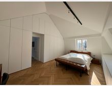 Modernes Schlafzimmer mit weißen Wänden und eingebauten Schränken, schräger Decke und einem großen Fenster, das Tageslicht hereinlässt. Der Raum enthält ein Doppelbett mit einer braunen Polsterkopflehne und eine Parkettboden in Fischgrätenmuster.