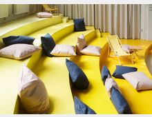 Gelber, abgestufter Sitzbereich mit geschwungenen Podeststufen, dekoriert mit zahlreichen Kissen in Pastell- und Dunkelblau-Tönen; im Hintergrund eine gelbe Liegestuhl-Sitzgruppe vor beigen Vorhängen.