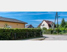 Zweifamilienhäuser in einer ruhigen Wohngegend mit Vorgärten, Hecken und Zäunen; gepflasterte Straßen im Vordergrund und ein Strommast mit oberirdischen Leitungen. Ein Straßenschild kennzeichnet eine Kreuzung.