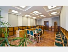 Ein moderner Besprechungsraum mit Holzvertäfelung, einem großen Tisch mit grünen Stühlen und einem Schreibtisch mit Computer. Die Decke ist mit quadratischen, eingebauten Leuchten gestaltet, und der Raum verfügt über einen Holzboden sowie mehrere Sitzgelegenheiten an den Wänden.