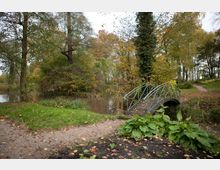 Ein Park mit einem kleinen Metallbogensteg, der über einen schmalen Bach führt. Umgeben von Bäumen mit Herbstlaub, Grasflächen, Kieswegen und dichter Vegetation.