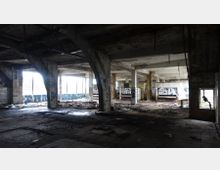 Innenraum einer verlassenen, verfallenen Fabrikhalle mit sichtbaren Betonstützen, großen Fenstern, bröckelnden Wänden und Graffiti. Der Boden ist mit Schutt und Trümmern übersät, während Tageslicht durch die Fenster fällt.