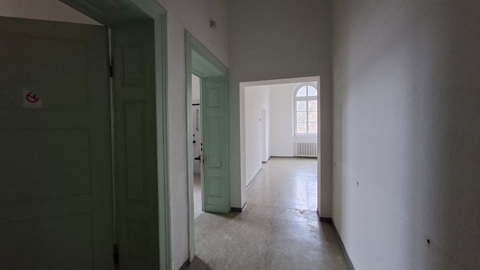 Schmaler, leerer Innenflur mit weißen Wänden, hellgrünen Holztüren und einem einfachen Bodenbelag. Am Ende öffnet sich der Gang zu einem hellen Raum mit großem Rundbogenfenster und einem Heizkörper darunter.