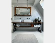 Modernes Badezimmer mit einer Waschtischkonsole aus Holz, darauf zwei rechteckige Waschbecken und ein großer Spiegel mit Holzrahmen. Die Rückwand besteht aus weißen Mosaikfliesen, und auf der Konsole befinden sich Pflanzen und Pflegeprodukte; helle Pendelleuchten hängen von der Decke.