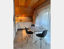 Kleine Küche mit schräger Holzdecke und Spotbeleuchtung, ausgestattet mit weißen Schränken und Arbeitsflächen. Im Vordergrund ein runder Esstisch mit drei schwarzen Stühlen, der auf einem hellen Steinboden steht, daneben ein großes Fenster mit weißen Vorhängen.