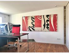 Ein modernes Büro mit Holzboden, einem schwarzen Schreibtisch, einem roten Bürostuhl und einem großen Kunstwerk an der Wand, das aus fragmentierten Teilen eines Coca-Cola-Logos besteht. Im Hintergrund steht ein Ventilator neben einem Fenster.