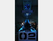 Ein dunkler Raum mit blauer Beleuchtung, in dem mehrere Personen an gegenüberliegenden Seiten eines Tisches an Computerstationen sitzen. Über den Arbeitsplätzen hängen industrielle Belüftungsgeräte mit erkennbaren Lüftern und der Aufschrift "R-42".