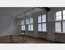 Ein schlichtes, leerstehendes Zimmer mit weiß gestrichenen Wänden, großen, hohen Fenstern mit Milchglas und geöffneten Flügeln sowie Heizkörpern unter den Fenstern. Im Raum steht ein einfacher, schmaler Holztisch auf einem Betonboden.