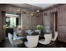 Esszimmer mit runden Esstisch und sechs hell gepolsterten Stühlen, umgeben von hölzernen Wänden in dunkler Farbe. Eine moderne, verzweigte Pendelleuchte hängt über dem Tisch, auf dem eine Glasvase mit Blumen und eine dekorative Schale stehen. Im Hintergrund ist eine offene Küche mit einem großen Fenster sichtbar.