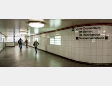 Unterführung einer S-Bahn-Station mit hell gefliesten Wänden, großen Deckenleuchten und Wegweiser zu verschiedenen Zielen, darunter "Oranienburg" und "Stresemannstraße". Einige Menschen gehen in Richtung eines Treppenaufgangs.