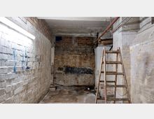 Ein leerer, heruntergekommener Kellerraum mit unverputzten Ziegelwänden, teils beschädigten und verschmutzten Oberflächen sowie sichtbaren Rohrleitungen unter der Decke. Eine alte Metallleiter steht rechts im Vordergrund, während auf der linken Wand Graffiti in Blau zu sehen sind.