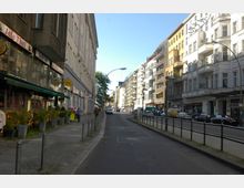 Straßenszene in einer Stadt mit mehrstöckigen Wohn- und Geschäftshäusern. Auf der linken Seite befinden sich Restaurants mit Außensitzbereichen, während auf der rechten Straßenseite Balkone und parkende Autos sichtbar sind; im Hintergrund führt die Straße in Richtung weiterer Gebäude.