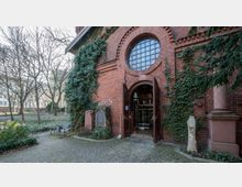 Backsteinfassade eines historischen Gebäudes mit einer offenen Holztür, die den Blick in das Innere freigibt. Die Wand ist von Efeu bewachsen, daneben stehen mehrere Grabsteine und eine Säule, umgeben von einem parkähnlichen Garten mit Bäumen und gepflastertem Weg.