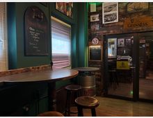Gemütliche Pub-Ecke mit hohen Holztischen und Barhockern neben einem Fenster mit Jalousien. Dunkelgrüne Wände und eine rustikale Holzvertäfelung mit Schiefertafeln, gerahmten Bildern und Guinness-Werbeschildern sowie einer Glastür zum Nebenraum.