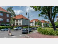 Straßenszene in einem Wohnviertel mit mehrstöckigen Häusern im Backstein- und Putzstil, roten Ziegeldächern und weißen Fenstern. Im Vordergrund geparkte Autos, eine ältere Frau mit Einkaufstrolley und ein Schild mit der Aufschrift „Winfriedstraße“.