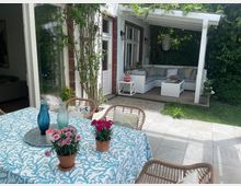 Ein überdachter Gartenbereich mit einer weißen Lounge-Sitzgruppe auf einer Holzterrasse, umgeben von Pflanzen. Im Vordergrund steht ein Gartentisch mit blauer Tischdecke, Blumen in Tontöpfen und dekorativen Glasvasen.