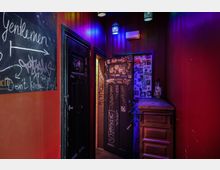 Enger Flur einer Bar oder eines Clubs mit roten Wänden, dekoriert mit buntem Neonlicht. Eine schwarze Tür und Wände sind mit Graffiti und Stickern bedeckt, daneben steht ein Holzschrank mit Flaschen.
