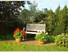 Holzbank auf einer gepflasterten Fläche in einem Garten, umgeben von dichtem Grün und blühenden Pflanzen. Zwei Blumentöpfe mit roten und weißen Geranien stehen links und rechts vor der Bank.