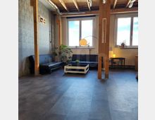 Ein gemütlicher Aufenthaltsraum mit industriellem Flair, ausgestattet mit schwarzen Sofas, einem weißen Couchtisch und Holzbalken, die die offene Decke mit sichtbaren Rohren stützen. Große Fenster sorgen für viel Tageslicht, und Pflanzen sowie eine Stehlampe ergänzen die Einrichtung.