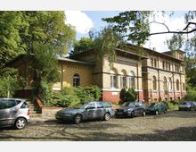 Ziegelstein-Gebäude mit historischen Rundbogenfenstern, umgeben von Bäumen und Grünanlagen. Im Vordergrund parken mehrere Autos auf einer gepflasterten Straße.