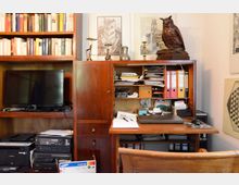 Ein Arbeitszimmer mit einem braunen Holzmöbel-Set, bestehend aus einem Regal mit Büchern, einem Schreibtisch mit Bürobedarf und Ordnern, sowie einem Fernsehgerät und Drucker. Auf dem Regal steht eine Eulenfigur aus Holz, und an der Wand hängen gerahmte Kunstwerke.