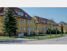 Mehrfamilienhäuser mit gelber Fassade und roten Ziegeldächern in einer Wohnsiedlung, umgeben von jungen Bäumen, Grünflächen und einem Gehweg. Ein Straßenschild mit der Aufschrift „Friedrich-Ebert-Ring“ ist sichtbar.