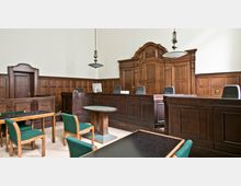 Historischer Gerichtssaal mit hölzernem Wand- und Mobiliarverkleidung, einer erhöhten Richterbank im Hintergrund und mehreren Tischen und Stühlen mit grüner Polsterung im Vordergrund. Zwei große Holztüren und hängende Lampen ergänzen die klassische Ausstattung.