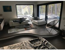 Modernes Wohnzimmer mit heller Eckcouch auf einem Podest, kleinem Rundtisch und Teppich vor großen Fenstern und einer Terrassentür mit Jalousien. Daneben stehen ein geflochtener Lounge-Sessel und eine Zimmerpflanze; draußen ist eine Holzterrasse mit Blick ins Grüne zu sehen.