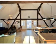 Wohnzimmer in einem Dachgeschoss mit sichtbaren Holzbalken, großen Fenstern mit weißen Rahmen, einem Esstisch mit Stühlen und einer Sitzecke mit Sofa, Sesseln und einem Couchtisch. Holzboden und moderne Lampen prägen den Raum.