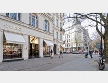 Straßenszene in einer Einkaufsstraße mit einem luxuriösen Gucci-Geschäft in einem historischen Gebäude. Links sind große Schaufenster mit weißen Markisen, rechts im Hintergrund weitere elegante Architektur und geparkte Autos.