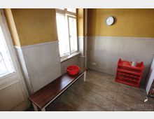 Ein einfacher Raum mit beige-gelben Wänden und einer halbhohen weißen Wandverkleidung. Links befindet sich eine Holzbank mit einer roten Schüssel darauf, an der Wand hängt eine runde Uhr, und rechts steht ein rotes Regal.