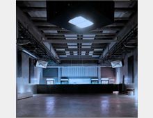 Großer Innenraum einer modernen Veranstaltungshalle mit minimalistischem Design. Zu sehen sind eine leere Tanzfläche, eine dunkle Bartheke im Hintergrund, symmetrisch angeordnete Leuchtkörper an der Decke sowie strukturelle Träger und Lautsprecher.