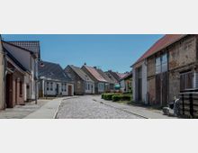 Gepflasterte Dorfstraße mit kleinen, einstöckigen Häusern, die teils renoviert und teils älter wirken. Die Gebäude haben Satteldächer, Gartenabschnitte und stehen entlang von Gehwegen unter blauem Himmel.