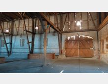 Innenraum einer rustikalen Scheune mit sichtbaren Holzbalken und Backsteinwänden. Im Vordergrund sind Holzkonstruktionen und einfache Holzmöbel zu sehen, im Hintergrund eine große Holztür mit Lichtdurchlässen und eine kleine Fensteröffnung an der rechten Wand.