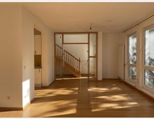 Helles, unvermöbliertes Wohnzimmer mit Parkettboden, großen bodentiefen Fenstern auf der rechten Seite und einer Holztreppe im Hintergrund hinter einer Glastür. Links neben der Glastür ist ein kleiner Küchenbereich zu sehen.