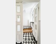 Detailaufnahme eines hellen Innenraums mit schwarz-weißem Schachbrettmuster auf dem Boden. Im Vordergrund sind geöffnete weiße Türen mit mehreren verzierten Fenstereinsätzen zu sehen, im Hintergrund führen Holztreppenstufen in einen leicht erhöhten Bereich mit einem Heizkörper an der Wand.