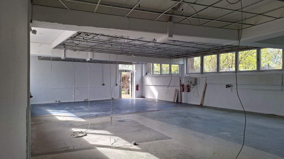 Großer, leerer Innenraum einer ehemaligen Werkstatt oder Lagerhalle mit Betonboden, weißen Wänden und abgehängter Decke im Umbau, aus der Kabel herabhängen. Entlang der rechten Seite befindet sich eine Reihe hoher Fenster mit Blick auf Bäume; hinten ist eine offene Tür nach außen sichtbar.