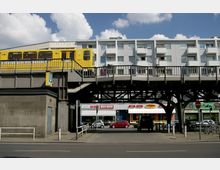 Gelbe U-Bahn auf einer erhöhten Stahlkonstruktion über einer Straße, darunter Geschäfte, parkende Autos und Wohngebäude im Hintergrund. Ein „U“-Schild weist auf eine U-Bahn-Station hin.