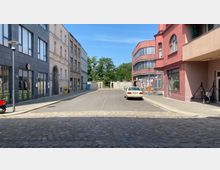 Pflasterstraße mit angrenzenden Gebäuden, darunter ein modernes graues Haus mit Schaufenstern und ein rosa Gebäude mit abgerundeten Ecken. Im Hintergrund eine Mauer mit einem Durchgang und dahinter Bäume.