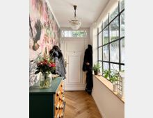 Flur mit heller Holztür und Oberlicht sowie großer Fensterfront rechts, die viel Tageslicht hereinlässt. Auf der linken Seite befindet sich eine grüne Holzkommode mit Blumenstrauß obenauf, daneben eine farbenfrohe Tapete mit Vogelmotiven; an der Türseite hängen mehrere Jacken an Haken. Holzboden im Fischgrätenmuster und ein Kronleuchter an der Decke komplettieren das Interieur.