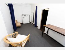 Ein schlichter Innenraum mit grauem Teppichboden, weißen Wänden und blauen Vorhängen. Der Raum enthält einen Kleiderständer, einen Spiegel, einen kleinen Holz- und Metalltisch sowie Sitzmöbel, darunter einen Korbstuhl mit Kissen und einen gepolsterten Stuhl.