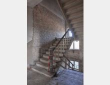 Ansicht eines alten, verfallenen Treppenhauses mit freiliegenden Ziegelwänden und verwitterten hölzernen Treppenstufen. Im Hintergrund sind große Fenster zu sehen, die diffuses Tageslicht in den Raum werfen.