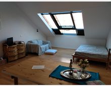 Dachgeschosszimmer mit Holzboden, einem großen Dachfenster, einem Einzelbett, einem kleinen Sofa mit Decke, einem Korbmöbel mit Fernseher sowie einem Holztisch mit Dekorationen wie Blumen und einem Silbertablett.