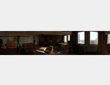 Panoramablick in einen großzügigen, loftartigen Raum mit abgenutzten Wänden, großen Fenstern mit Blick auf Hochhäuser und einer Mischung aus Wohn- und Musikstudioeinrichtung. Zu sehen sind Sofa und Couchtische, ein Arbeitsbereich mit Technik sowie mehrere Gitarren und Verstärker entlang der Fensterfront.