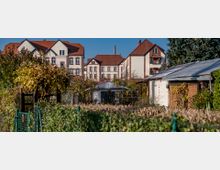 Mehrere Reihenhäuser mit roten Dächern und heller Fassade im Hintergrund, davor eine Kleingartenanlage mit Lauben, Schuppen und dichten Hecken, umgeben von Bäumen und Sträuchern.