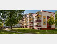 Mehrstöckiges Wohngebäude mit gelb-weißer Fassade und roten Balkonen, umgeben von einer grünen Wiese mit Bäumen und Gehwegen. Im Vordergrund ist ein großer Baum, während im Hintergrund Spielplatzstrukturen und weitere Gebäude zu sehen sind.