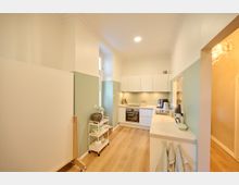 Helle, moderne Küche mit weißen Schränken, hochglänzender Arbeitsfläche und Elektrogeräten wie Backofen und Kaffeemaschine. Die Wände sind zur Hälfte in Grün gestrichen, der Boden besteht aus hellem Holz, und es gibt Zugang zu einem angrenzenden Raum durch einen offenen Durchgang.